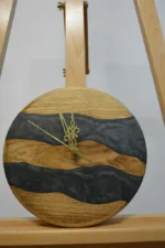 Tömör fa falióra epoxigyantával (23 cm), két antracit színű gyanta folyóval, arany mutatókkal, szemből fotózva állványon.