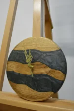 Tömör fa falióra epoxigyantával (23 cm) antracit színben, arany mutatókkal, ferde nézetben fa állványon.