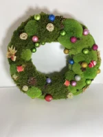 Színes karácsonyi gömbökkel, tobozokkal és szalmadíszekkel díszített mohakoszorú fehér háttér előtt.