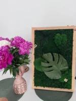 Mohakép Monstera levéllel fa keretben, mellette egy váza rózsaszín pünkösdi rózsákkal.