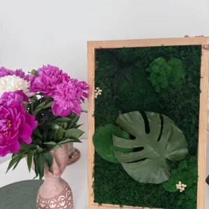 Mohakép Monstera levéllel fa keretben, mellette egy váza rózsaszín pünkösdi rózsákkal.